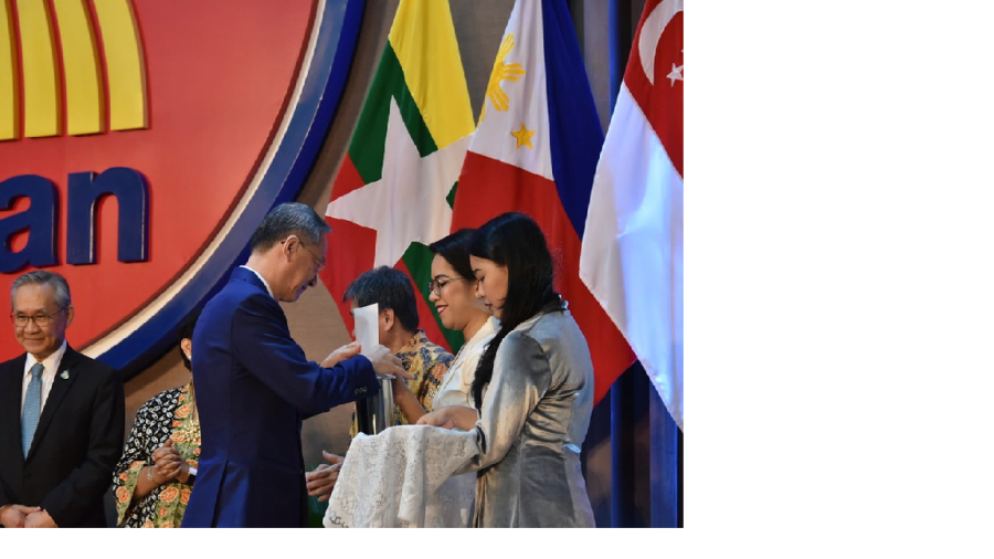 Man in suit hands papers to woman before flags of Myanmar, Philippines, and Singapore.
